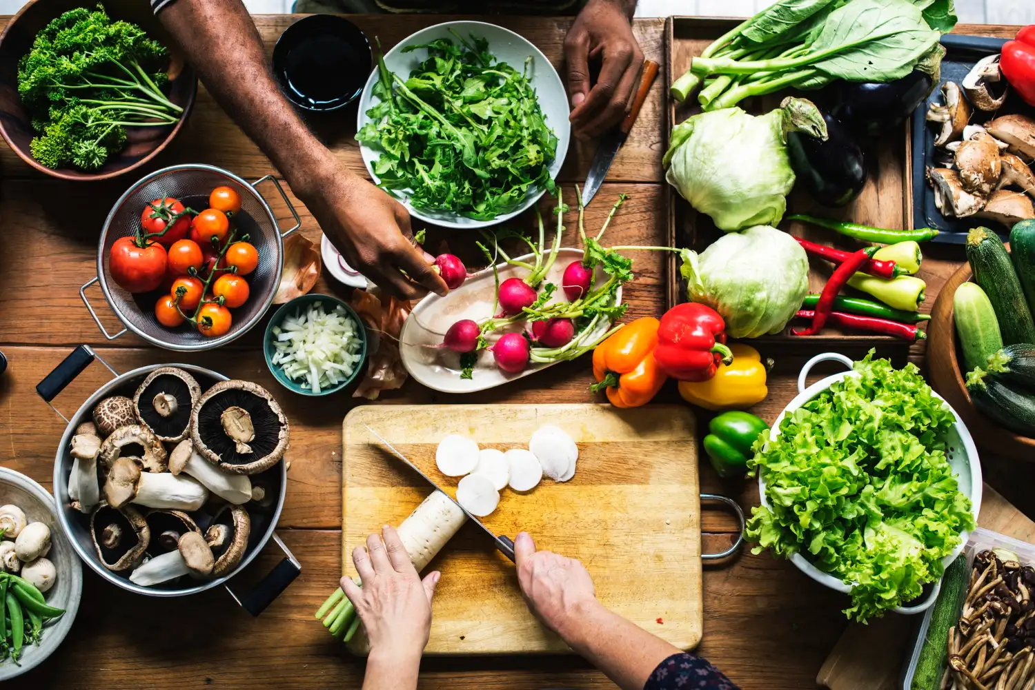 Pessoas preparando uma variedade de vegetais frescos sobre uma mesa de madeira, com folhas, tomates, rabanetes, pimentões, cogumelos e outros ingredientes.