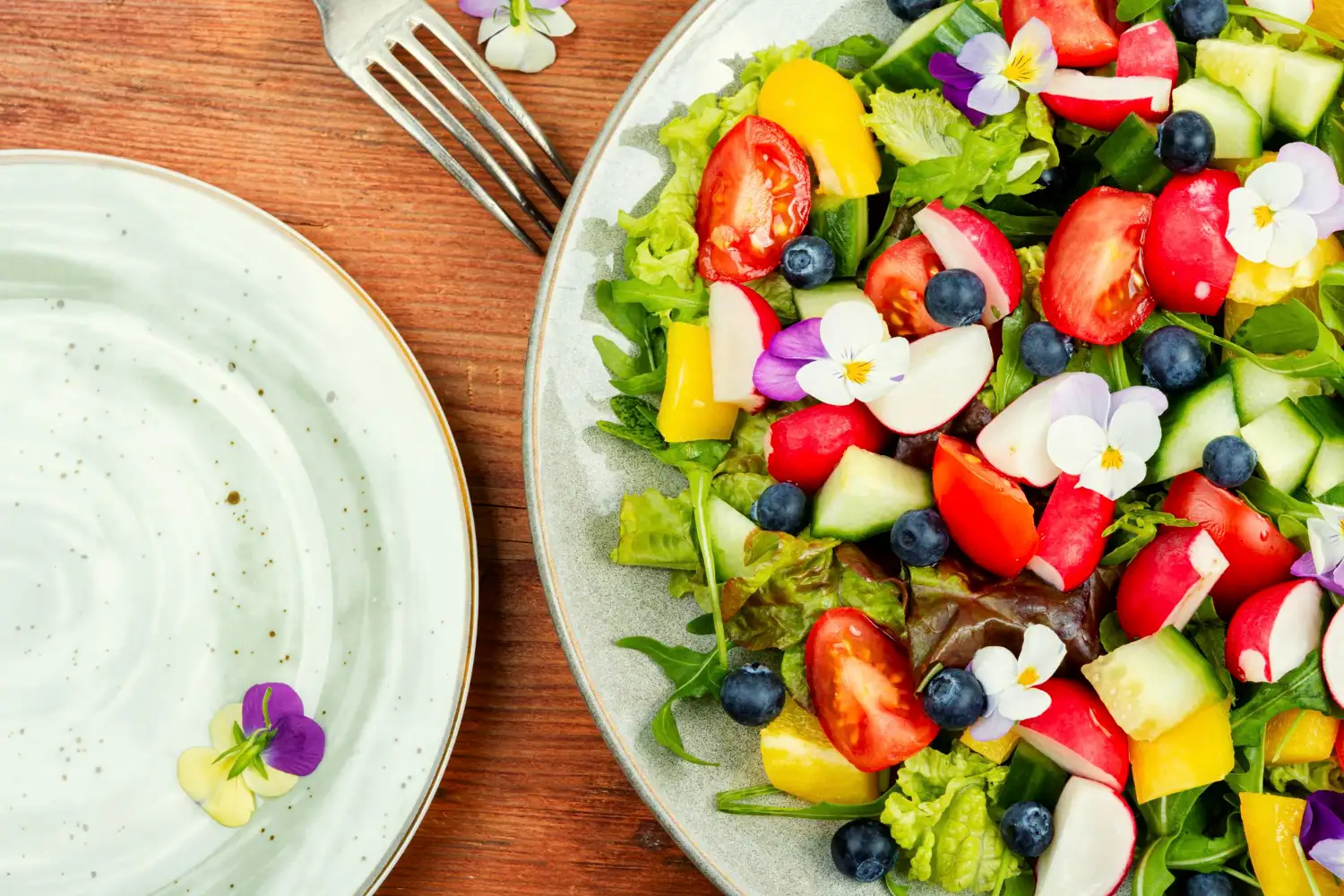 29309 Salada colorida com folhas verdes, tomate, pepino, rabanete, mirtilos, pimentão amarelo e flores comestíveis servida em prato sobre mesa de madeira.